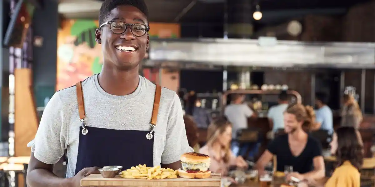 Portrait Of Young Waiter Serving Food To Customers In Busy Bar Restaurant