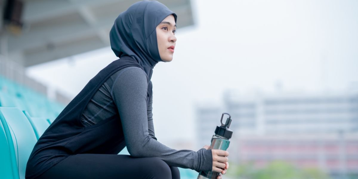 A young arab-american woman wearing a black hijab is doing exercise and resting tired at the outdoor stadium in the morning.