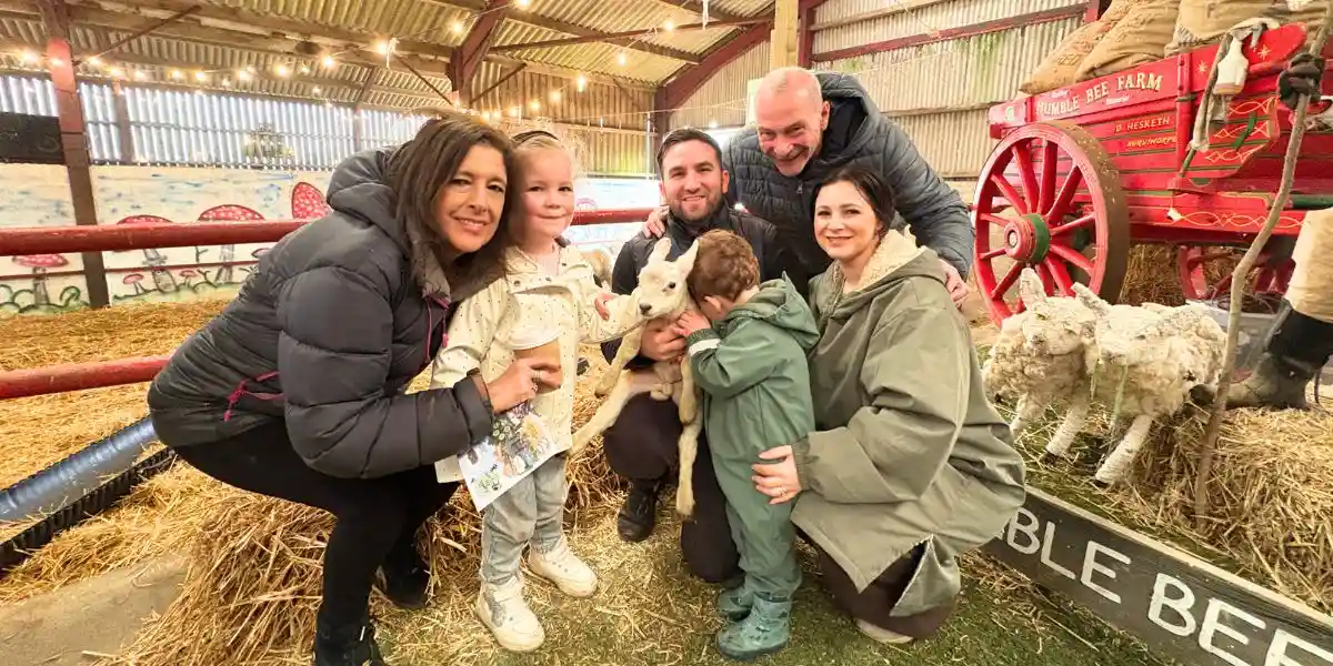 family cuddling farmed lambs with hay bales and a red tractor in background
