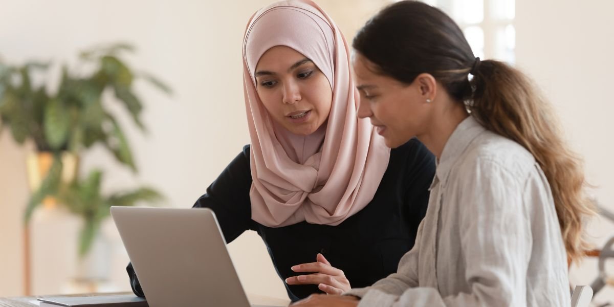 Concentrated young arabian women in hijab sitting with smiling colleague at table, looking at computer screen, explaining new company software.