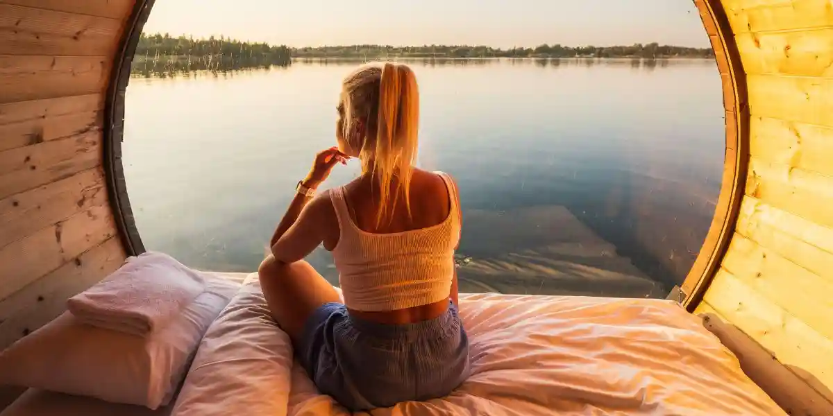 woman relaxing on a cozy bed inside a modern wooden barrel house, looking at a calm lake during sunset.