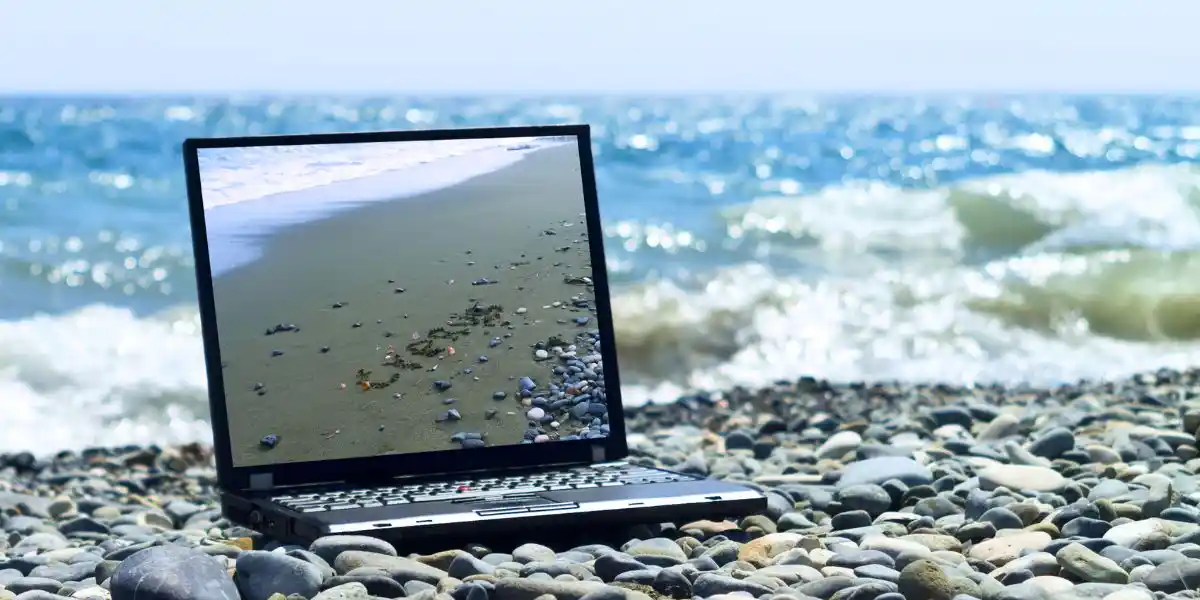 Laptop with picture of Cyprus beach on its monitor laying on gray pebbles in front of blue wavy sea